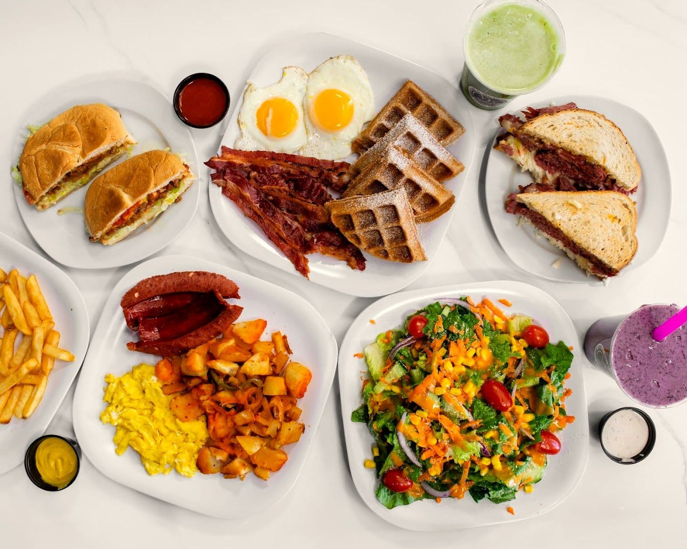 A spread of NYC Style Deli plates: breakfast sandwiches, eggs and bacon, pastrami on rye, fries, salad and smoothies on a marble counter